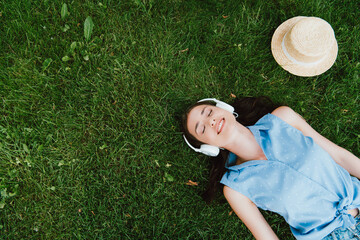 top view of cheerful woman lying on grass and listening music near straw hat