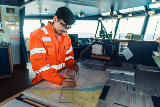 Filipino deck Officer on bridge of vessel or ship wearing coverall during navigaton watch at sea . He is plotting position on chart