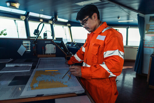 Filipino deck Officer on bridge of vessel or ship wearing coverall during navigaton watch at sea . He is plotting position on chart