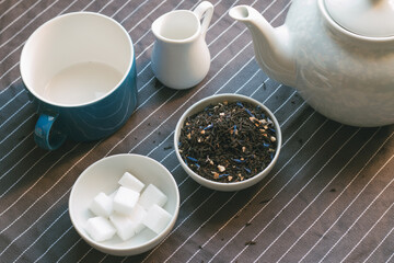 Tea set on a striped grey cloth. Tea pot, bowl with dried leaves, bowl with sugar cubes, cup en little milk jug