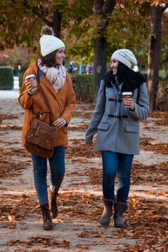 Best friends chatting in the park in the fall with their coffee to go. They're wearing a casual coat and a wool hat.