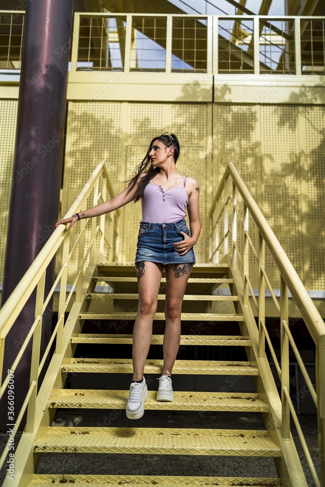 Poster Young rebellious caucasian female with tattoos and in jeans skirt posing on the yellow stairs