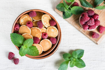 Trendy food pancake cereal, mini pancakes in a bowl with fresh raspberries and mint on a light textile background, top view