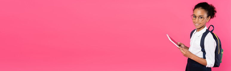 smiling african american schoolgirl in glasses with backpack using digital tablet isolated on pink,...