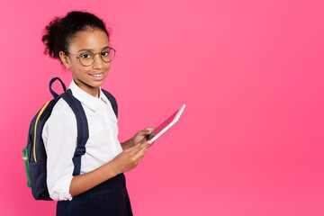smiling african american schoolgirl in glasses with backpack using digital tablet isolated on pink