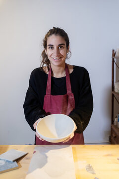 Smiling young ceramist woman showing ceramic bowl in pottery workshop
