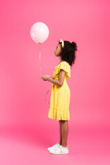 side view of curly african american child in yellow outfit with balloon on pink background