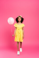 Obraz na płótnie Canvas full length view of smiling curly african american child in yellow outfit with balloon on pink background