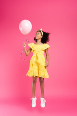 full length view of smiling curly african american child in yellow outfit flying with balloon on pink background
