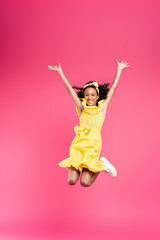 full length view of happy curly african american child in yellow outfit jumping with hands in air...