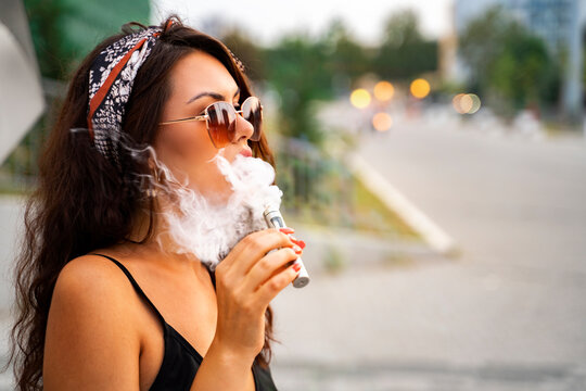Amazing, attractive boho brunette enjoying smoking e cigarette on city street  