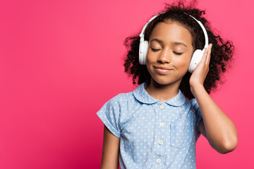 smiling curly african american kid in headphones with closed eyes isolated on pink
