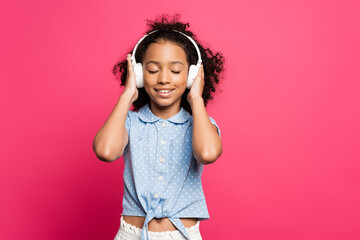 smiling curly african american kid in headphones with closed eyes isolated on pink