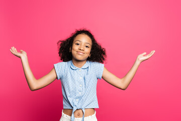 smiling cute curly african american kid showing shrug gesture isolated on pink