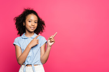 smiling cute curly african american kid pointing with  fingers aside isolated on pink