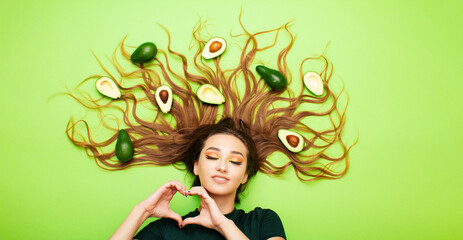 beautiful girl with avocado fruits on long hair, young woman shows shape of heart with hands,...