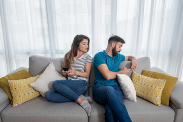 Obraz premium Young boyfriend making fun and jealous his girlfriend pretending that he is texting with other girl on his cell or mobile phone while they sitting on the sofa in their apartment