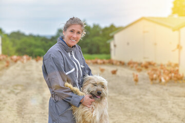 Portrait of a farmer petting her dog and standing in the middle of her chicken farm