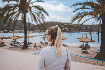 Fototapeta na wymiar A young blonde girl walking in the port of Soller in Palma de Mallorca Spain. Summer of 2020 Her blonde and light brown hair is half up with a ponytail. Palm trees, sun chairs and white sand, mountain