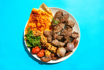 Famous traditional Arabic, Turkish, Israel food. Arabic grilled chicken, lamb, beef, sausages, onion, tomatoes, green, pita bread on white plate on blue background, top view. Mixed meat