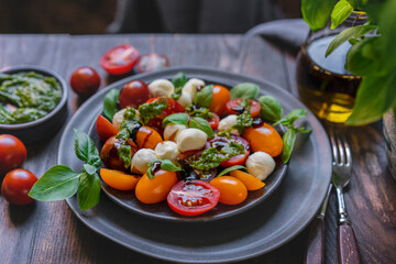 Caprese salad with fresh cherry tomatoes, mini mozzarella, pesto, basil, olive oil and balsamic sauce on blue plate, wooden rustic table. Mediterranean diet concept. Italian healthy food. Copy space.