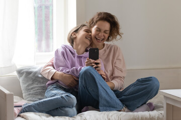 Fototapeta na wymiar Overjoyed mother and teenage daughter having fun with smartphone, relaxing on cozy bed at home, hugging, laughing at joke, happy excited mum and teenager girl using phone together, leisure time