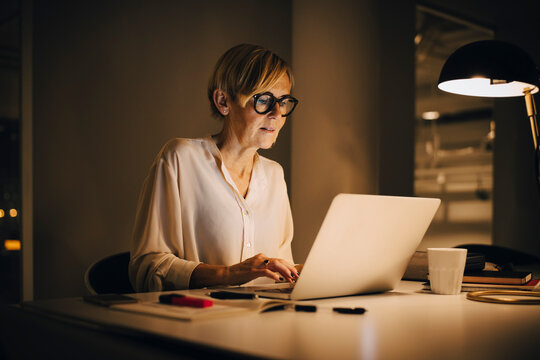 Determined businesswoman using laptop while working late in illuminated creative workplace