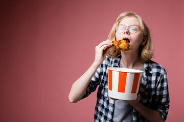 A swede girl in glasses and a plaid shirt raised a chicken leg to her mouth and holds a striped bucket in her hands with food on a pink background