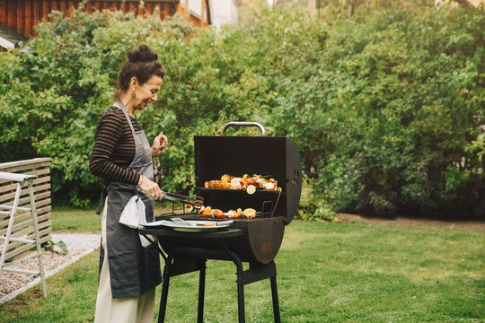 Side view of smiling woman cooking dinner on barbecue grill at back yard during garden party