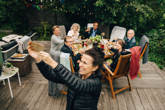 Smiling senior woman taking selfie with friends sitting at dining table in back yard during dinner party