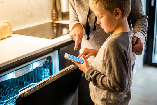 Boy learning from mother while using mobile app over dishwasher in kitchen at smart home