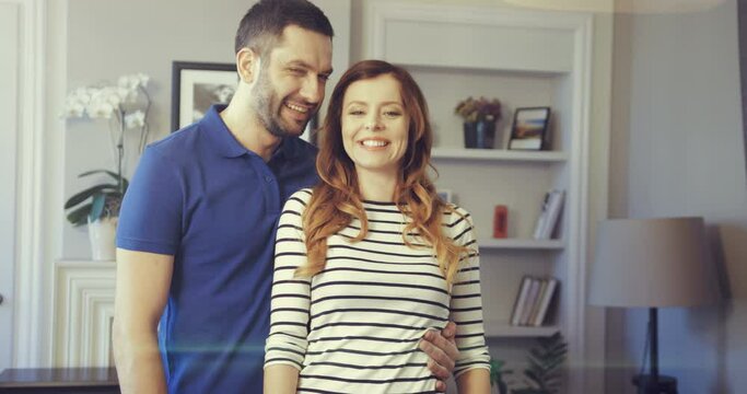 Portrait of happy couple standing in apartment and hugging, posing for camera. Husband carefully hugs his wife, wife looks at husband in love.