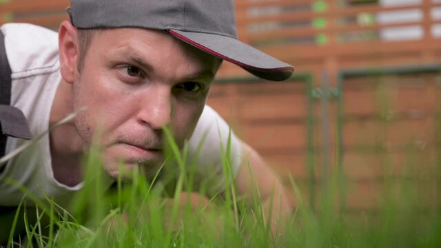 Crazy pedantic man in cap cutting green grass with scissors in private yard. Meticulous Male worker mowing high grass near house. Outdoor seasonal household works.