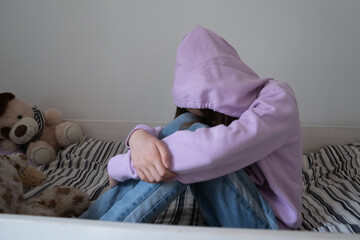 Lonely stressed teenage girl wearing hood sitting on bed alone, crying, thinking about troubles, hiding face, sad upset teenager feeling misunderstood and lonely, child and psychological problem