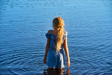 Alone child blond girl standing in the water in the light of the setting sun in summer