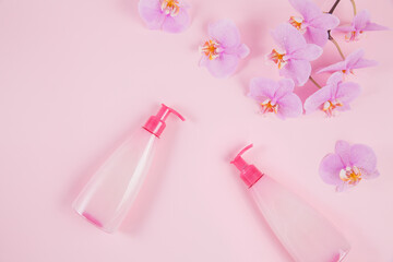 Two plastic dispenser bottles with liquide cosmetic soap, intimate wash or shower gel and pink orchid flowers on light purple background. Spa and women's hygiene concept