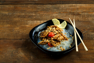 Nastol black plate with funchose and chicken with vegetables in pad Thai sauce next to chopsticks.