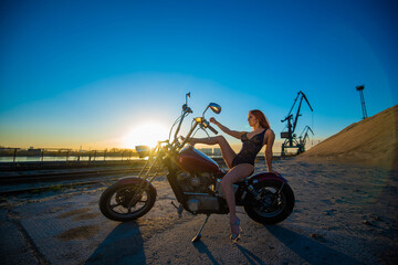 Young redhead woman in erotic lingerie in high heels sits on a motorcycle. Silhouette of a sexy girl on a bike. Industrial landscape.
