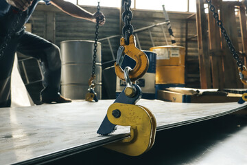 A worker in a factory lifts a bundle of sheet metal using a crane and mechanical clamps. Metal...
