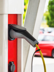 A charging cable for electric vehicles is plugged into a red-white charging station, cars are visible in the background.
