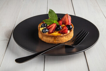 Fresh homemade fruit tart or cake with strawberries, mint leaves and blueberries on black plate which is on white wood, front view