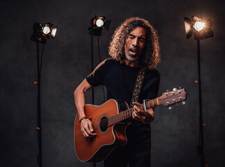 Middle aged hispanic musician in black t-shirt emotionally singing and playing guitar. View of musician in the spotlight