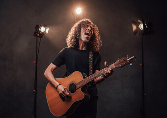 Middle aged hispanic musician in black t-shirt emotionally singing and playing guitar. View of musician in the spotlight