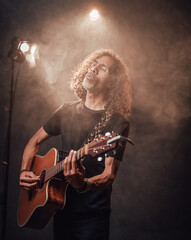 Hispanic musician in black t-shirt emotionally singing and playing guitar in stage lights, surrounded by smoke