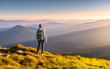 Fotobehang Oranje Mooie bergen in mist en staande jonge vrouw met rugzak op de piek bij zonsondergang in de zomer. Landschap met sportief meisje, groen gras, bos, heuvels, blauwe lucht met zonnestralen. Reizen en toerisme  © den-belitsky