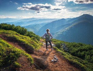 Beautiful mountains and standing young woman with backpack on the trail at sunset in summer....
