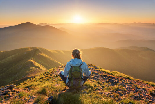 Young woman with backpack sitting on the mountain peak and beautiful mountains in fog at sunset in summer. Landscape with sporty girl, green forest, hills , sky, sunbeams. Travel and tourism. Yoga