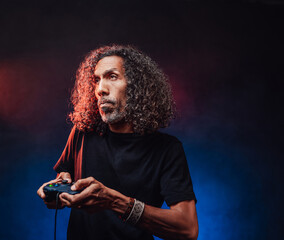 Middle aged hispanic male with long curly hair playing video game with joystick, looks focused. Hi-tech concept with vr glasses
