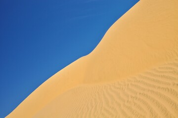 Sand dune against bright blue sky and sea. View on the sandy slope of the sand dune in hot sunny day. Sand dune surface close-up. Sand texture. Sand pattern full frame photography. Sand ripples.