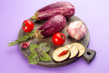 Summer vegetables on wooden board on purple background:ripe eggplants,tomatoes,fresh rosemary,purple basil leaves,garlic and hot pepper.Top view